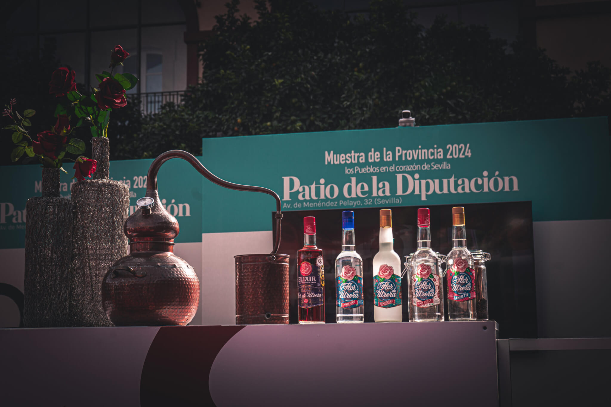 Botellas de Anís Flor de Utrera y licor Elixir junto a un alambique tradicional y decoración floral en el stand de la Feria de la Diputación de Sevilla 2024.