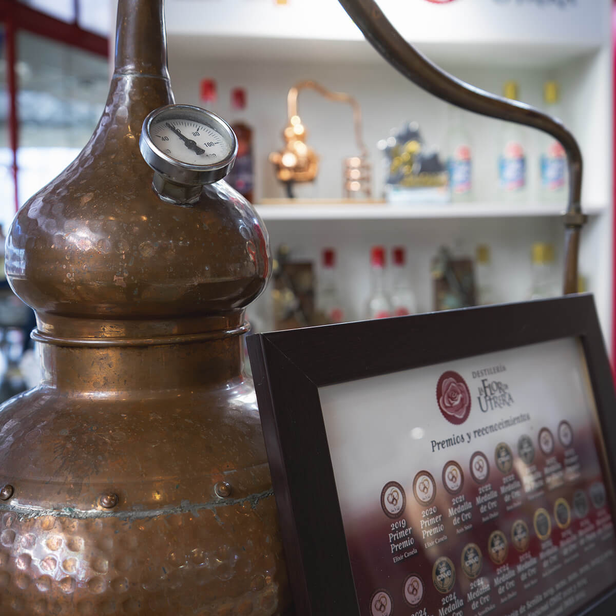 Detalle de alambique tradicional de cobre y panel de premios y reconocimientos de La Flor de Utrera, con botellas de anís al fondo durante el Salón H&T Málaga 2025.