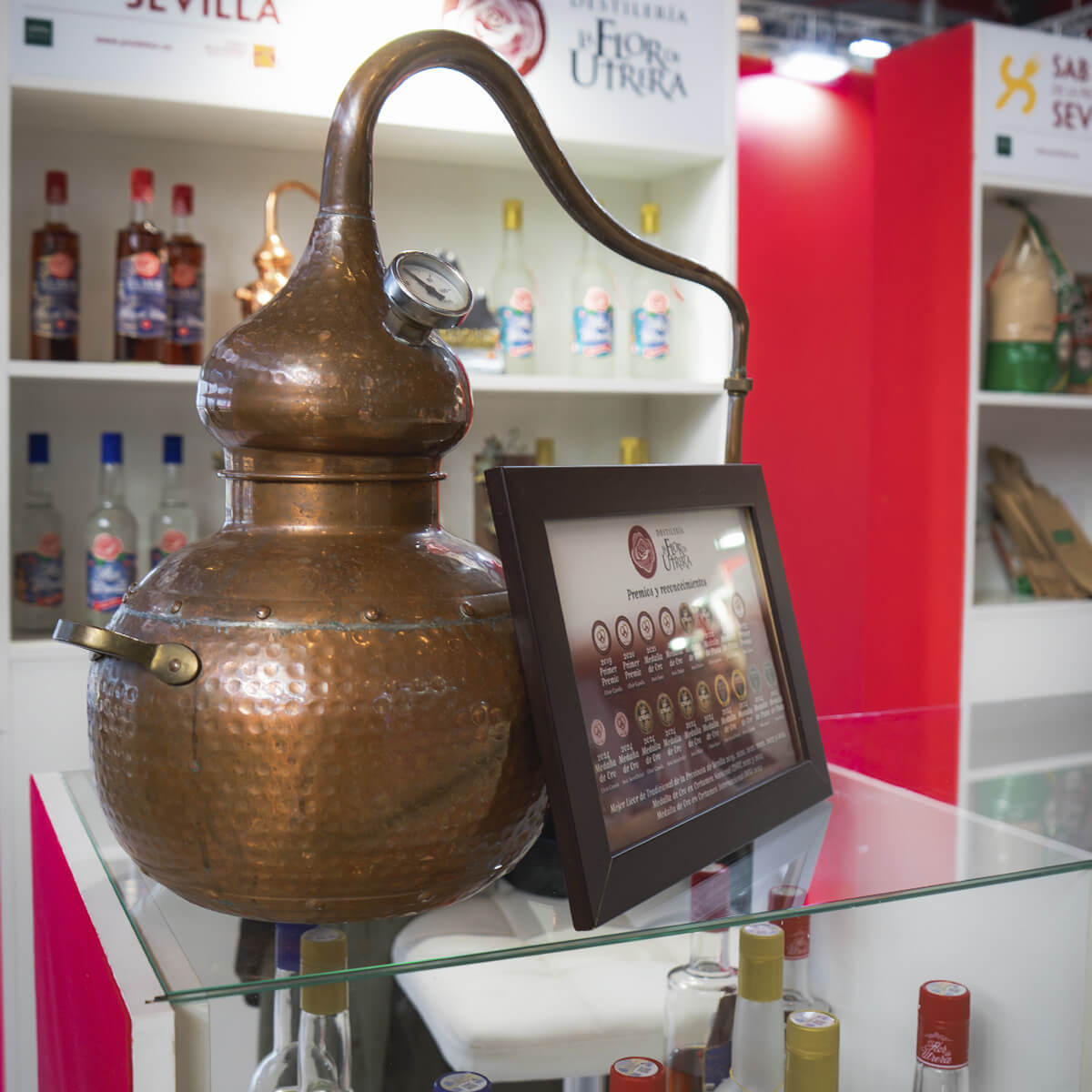 Primer plano del alambique de cobre tradicional utilizado en la destilación de anís, expuesto en el stand de La Flor de Utrera durante el Salón H&T Málaga 2025, junto a un cartel con los premios obtenidos por la destilería.